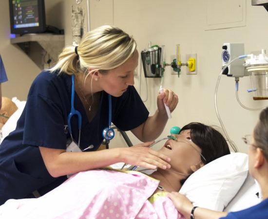 Nursing students in a lab simulation look over a mannequin in a hospital bed.
